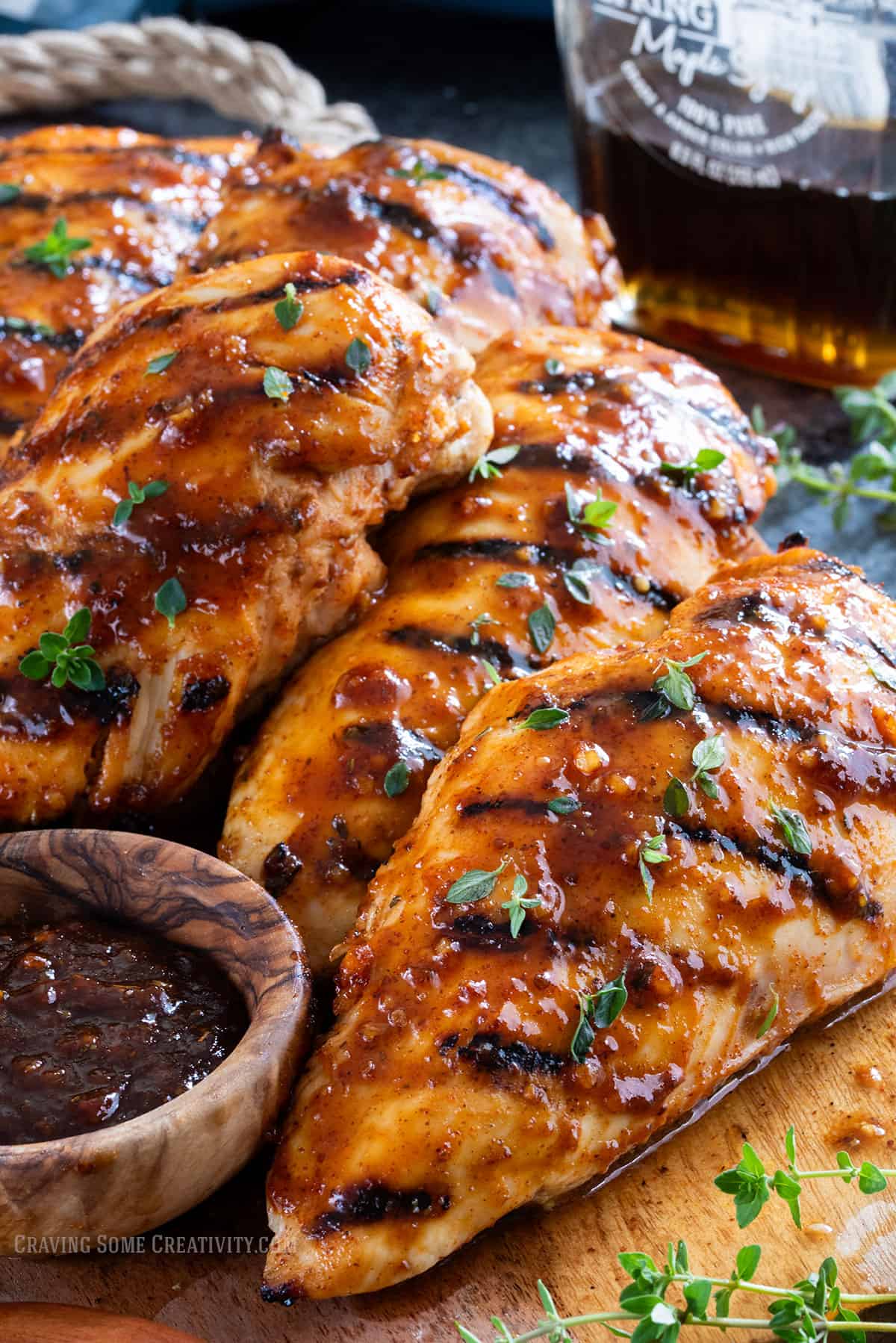 Several grilled chicken breasts with sauce on a wood board with a bottle of maple syrup.