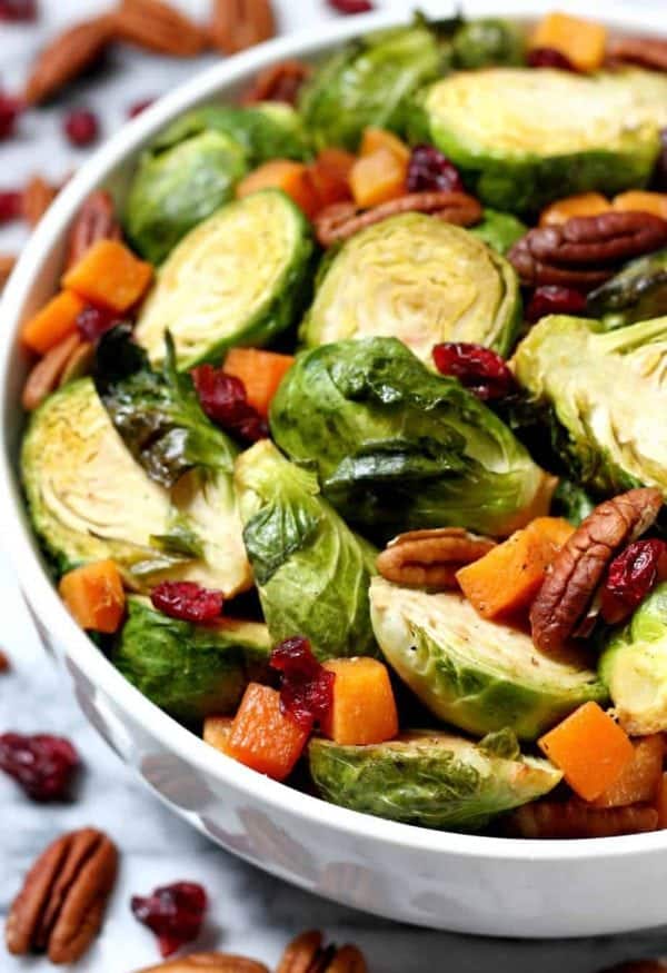 Maple roasted brussel sprouts with chopped squash, cranberries, and pecans in white bowl.