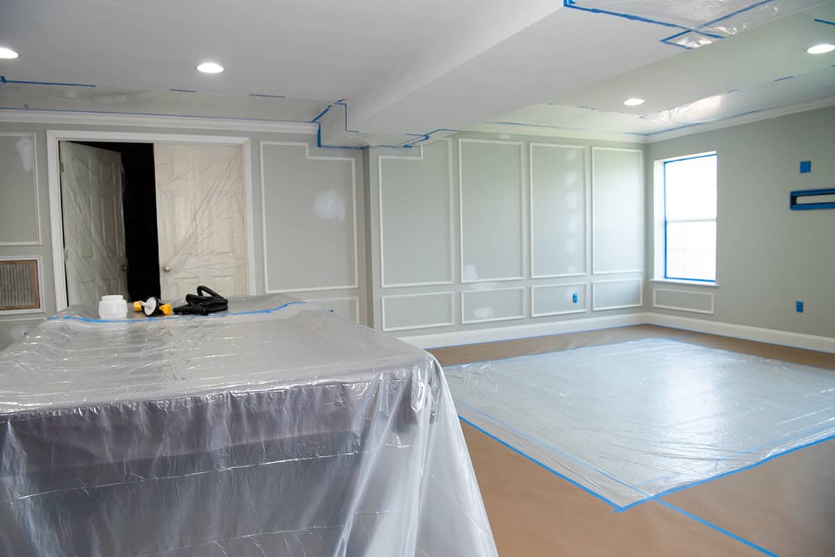 A basement prepped for painting with a sprayer with tarp on floors and masking on ceilings and furniture.