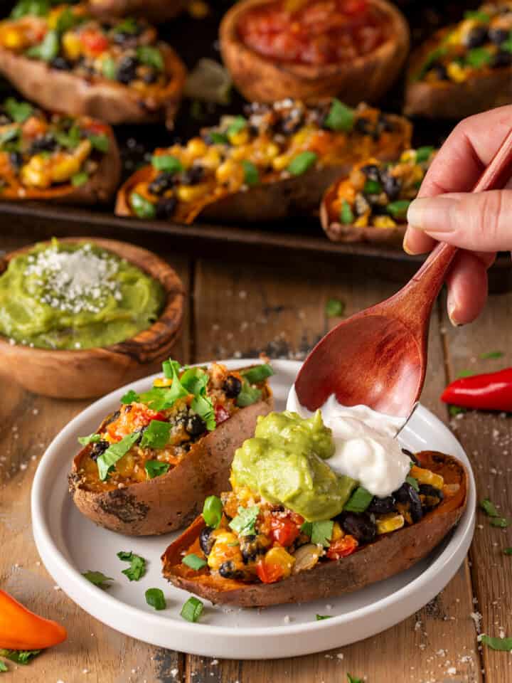 Two stuffed Mexican sweet potatoes in front while a hand tops them with guacamole and sour cream.