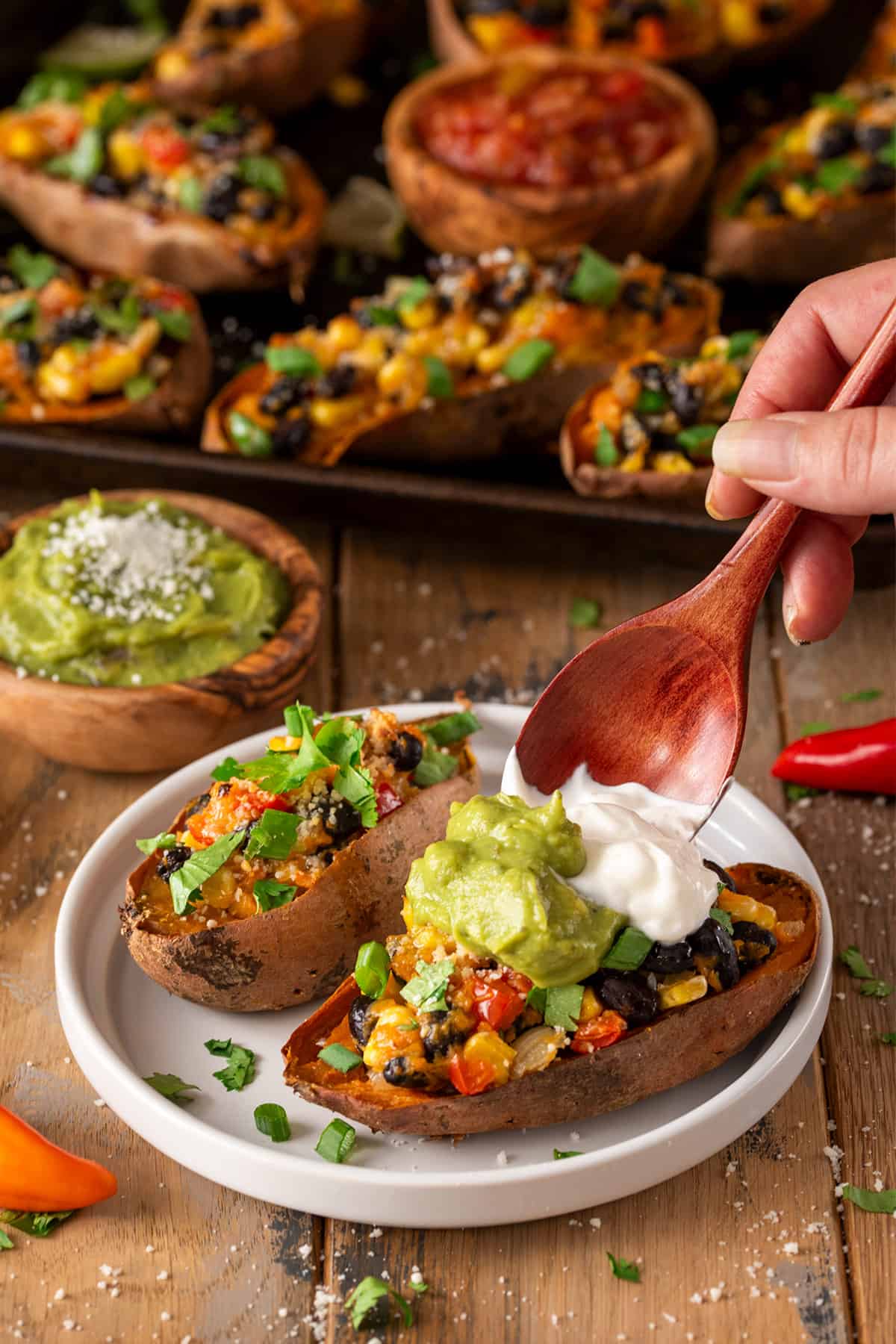 Two stuffed Mexican sweet potatoes in front while a hand tops them with guacamole and sour cream.