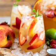 Two glasses of Moscato sangria with limes, apples, and peaches scattered around a serving tray.