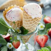 Mason jar with two waffle cones full of strawberry ice cream with strawberry blooms in foreground and empty cones and ice cream scoop in background.