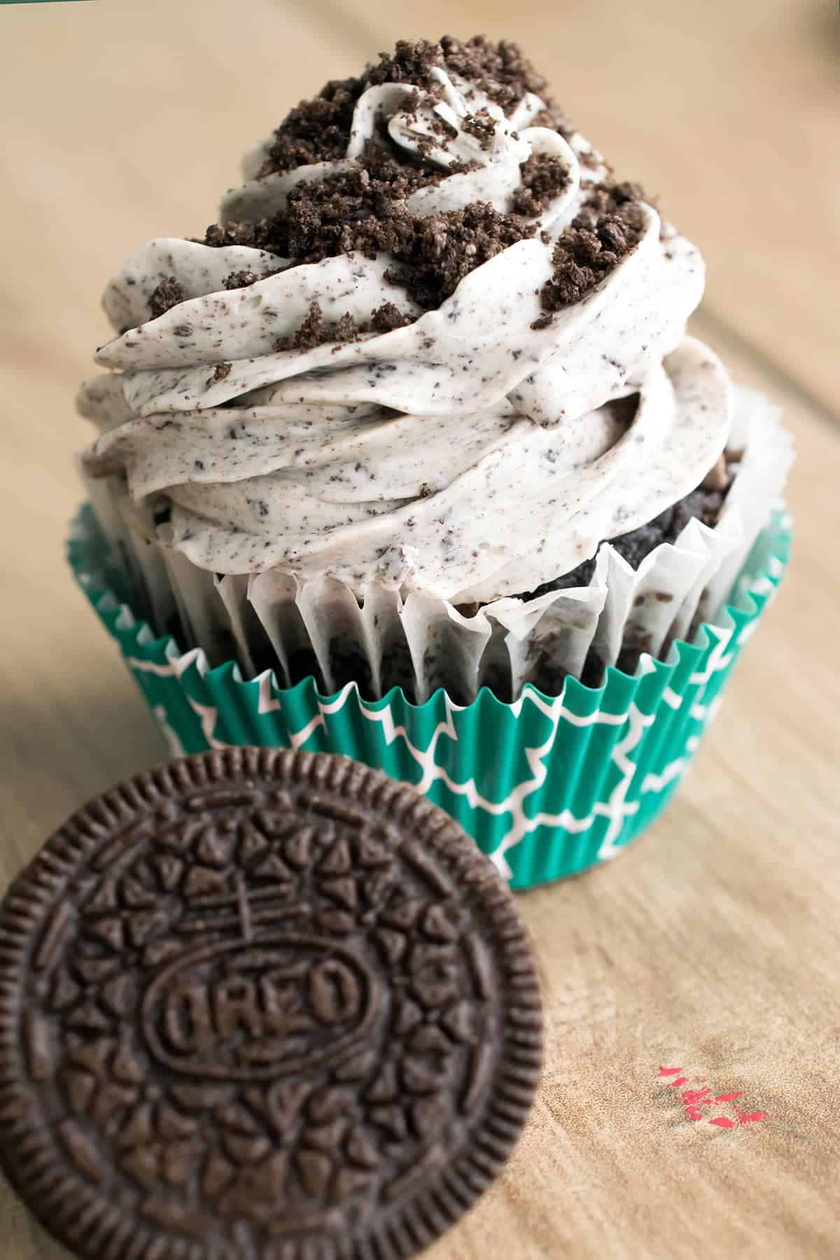 Cookies and Cream frosting swirled on chocolate cupcake, dusted with Oreo crumbles, in turquoise liner, beside whole Oreo cookie.