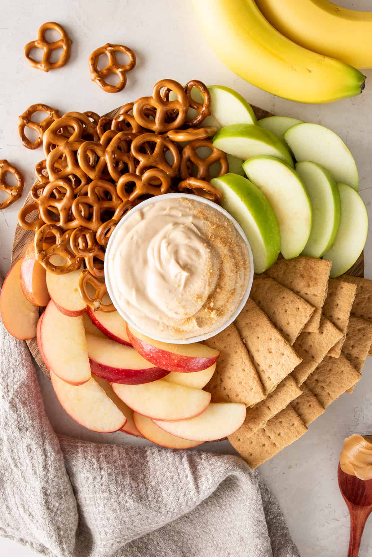 Overhead layout of peanut butter dip with apples, graham crackers, pretzels, and bananas spread around.