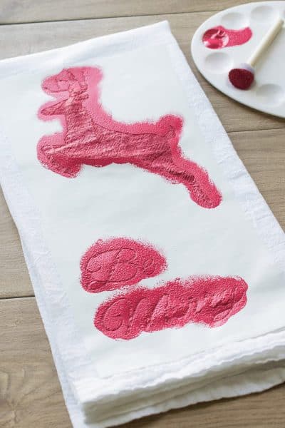 Deer and Be Merry message stencil on white tea towel with red paint and paint palette in background.