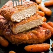Breaded baked pork chops with baby carrots. Top pork chop sliced in half with fork piercing through it.