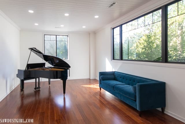 White plank drop-down ceilings with recessed lights in brightly lit great room with large windows, piano and turquoise couch.