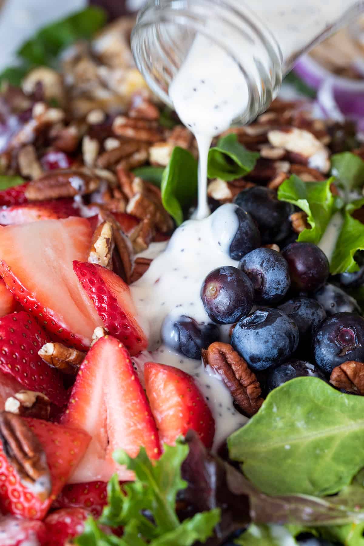 Homemade Poppyseed salad dressing recipe being poured over a mix of strawberries, blueberries, pecans, and lettuce greens.