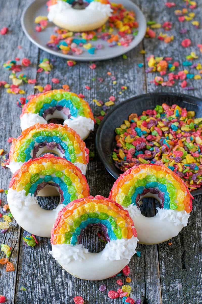 Homemade DIY rainbow donuts with Fruity Pebbles cereal rainbows and white glazed clouds on wood surface next to black bowl with cereal.