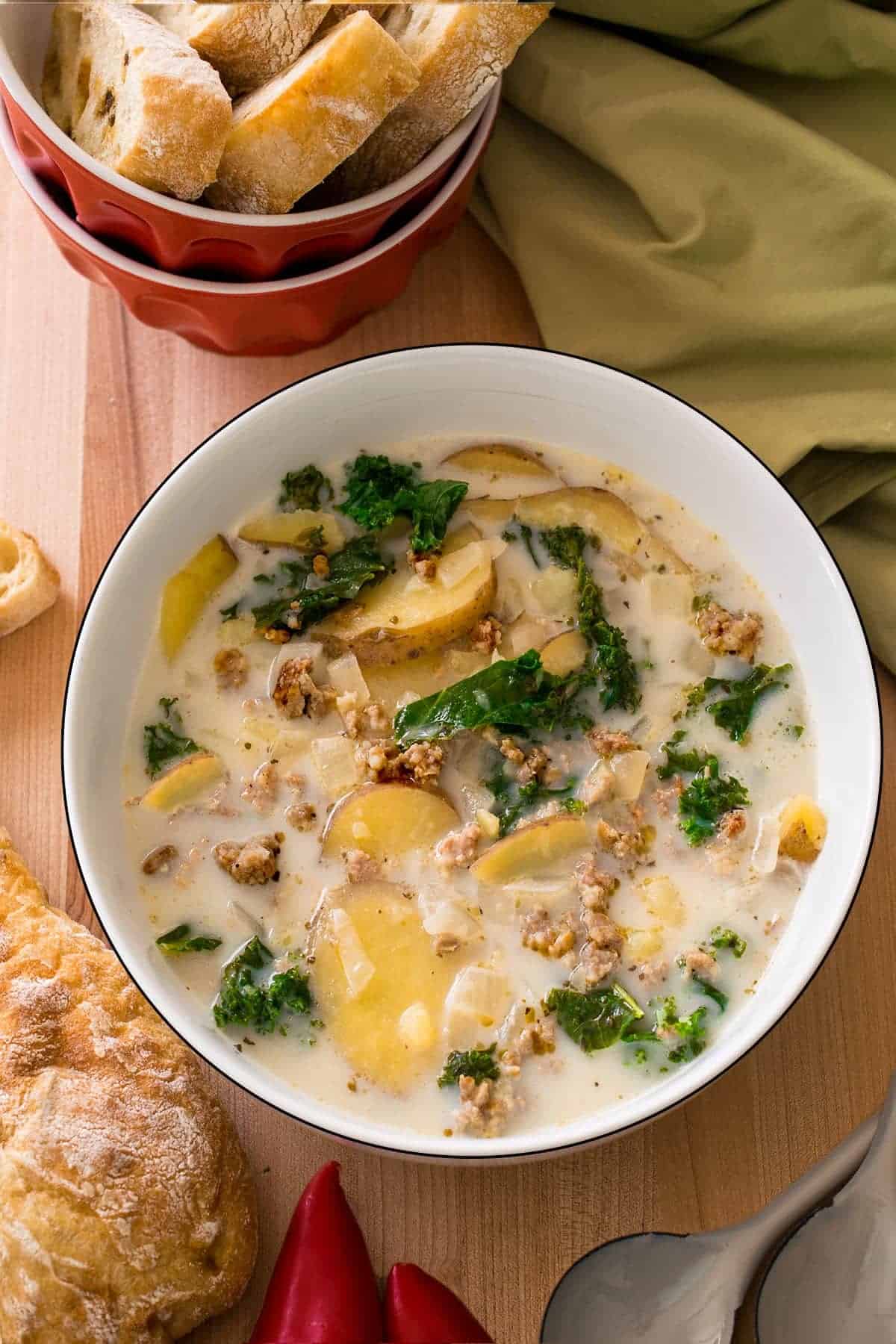 Italian sausage kale soup in white bowl on cutting board with crusty bread on the side and red bowl of crusty bread slices behind.