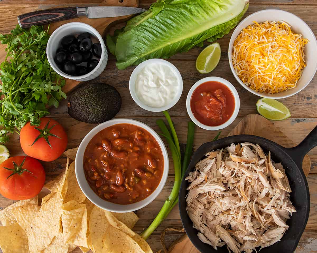 Layout of chicken chili nachos ingredients including salsa, sour cream, cheese, lettuce, tomato, olives, and avocado.