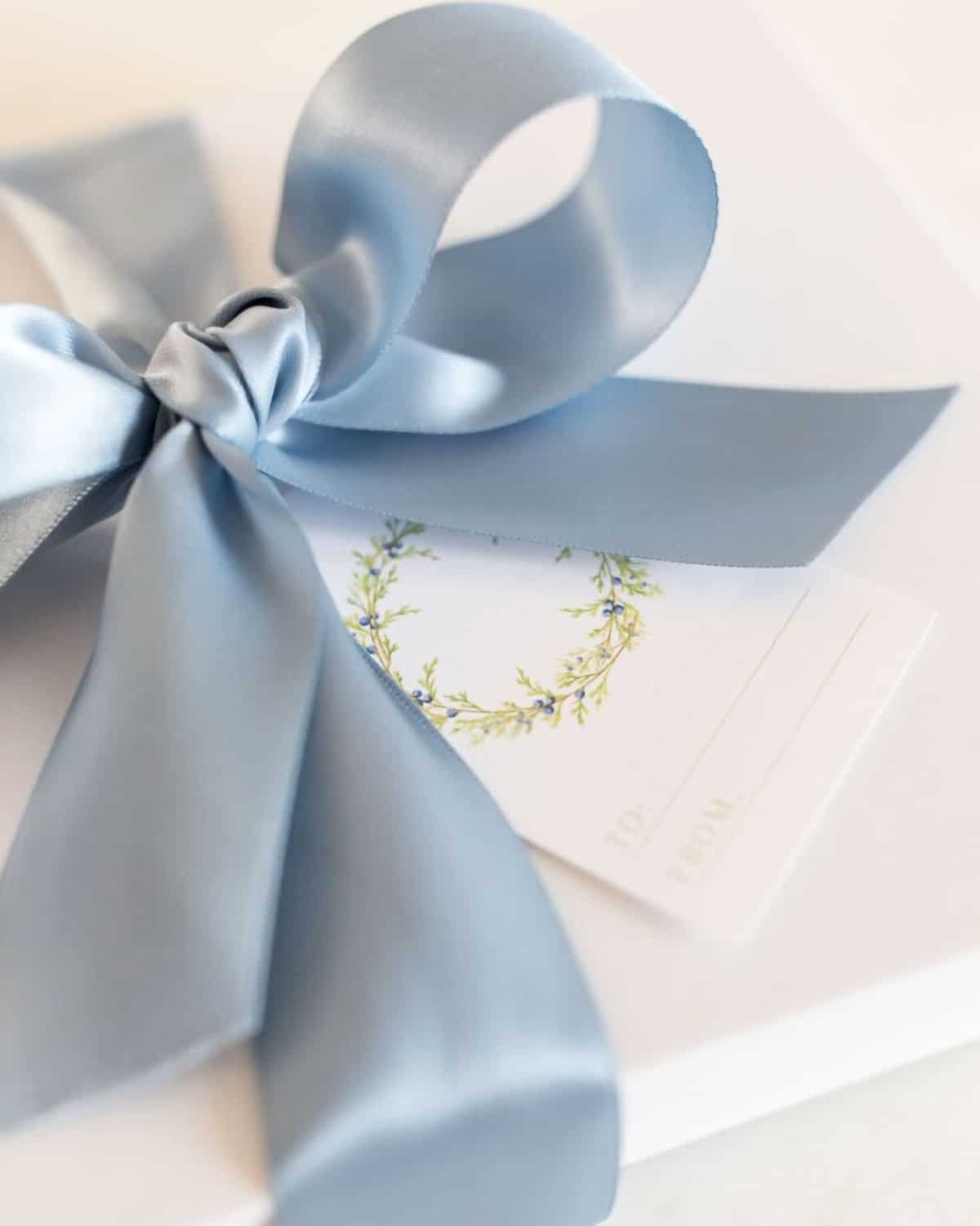 White covered Christmas package tied with large Robin blue bow qith Juniper wreath themed gift tag featuring blue berries.