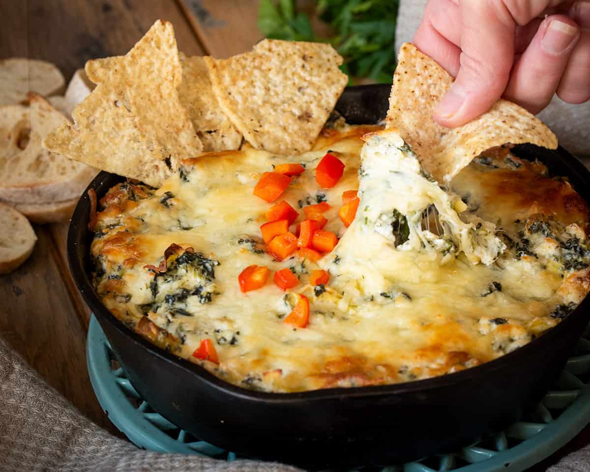 A hand dipping a tortilla chip in warm spinach dip and pulling away with a cheese pull.
