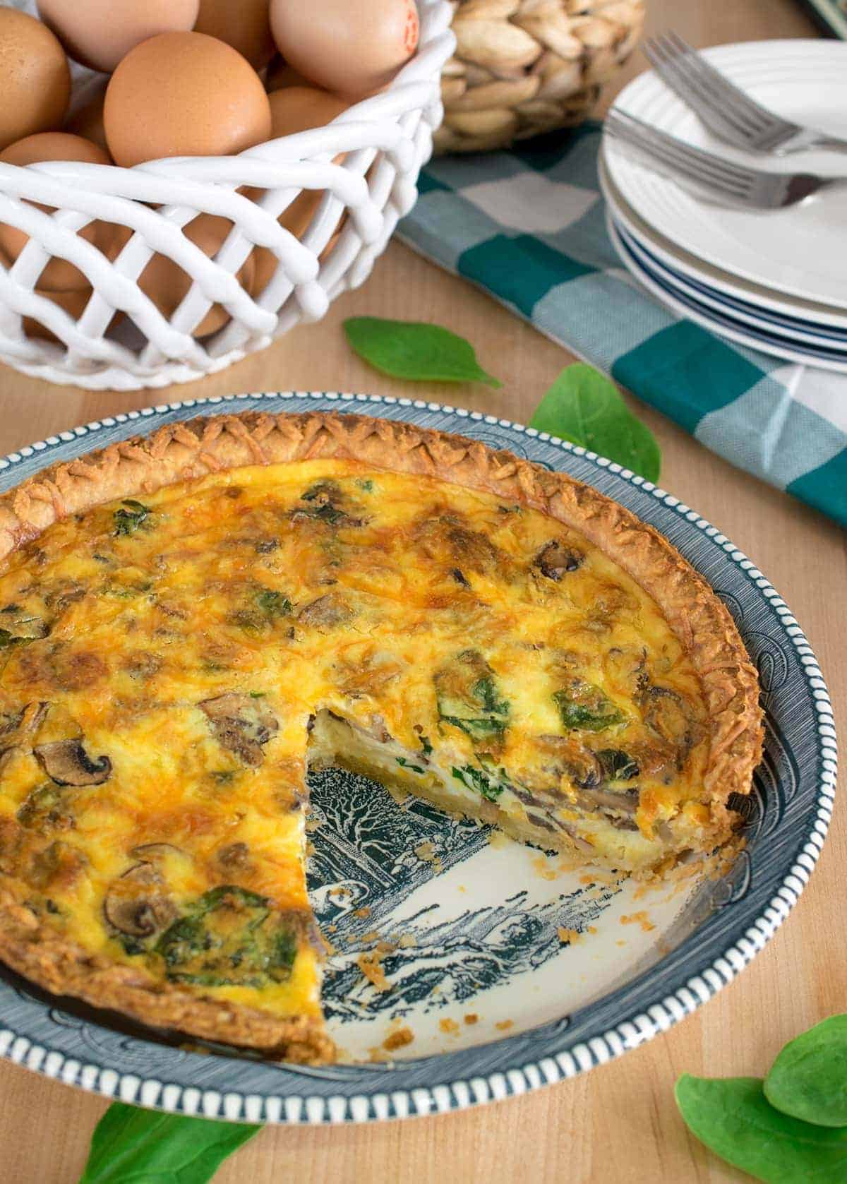 Spinach and mushroom quiche with crust in pie plate with one missing piece. Basket of eggs and stack of plates in background.