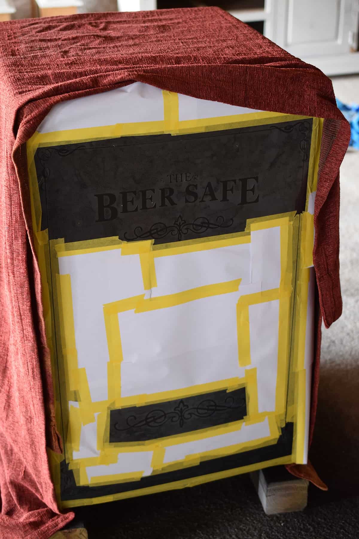 A painted fridge underneath a drop cloth with front door taped off in yellow tape and beer safe stencil applied. 