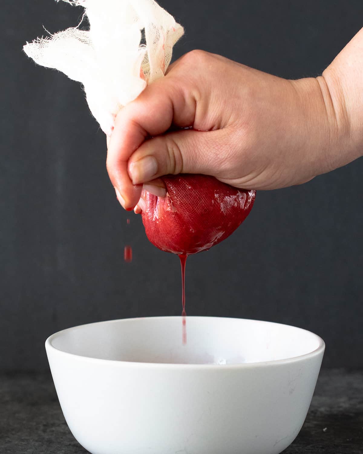 A hand squeezing strawberry puree through a cheesecloth sieve.