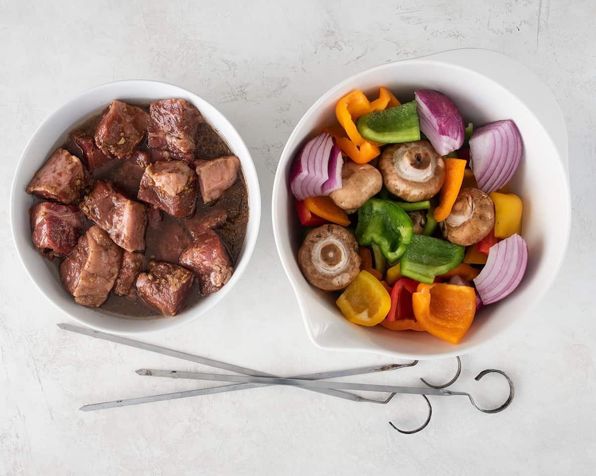 A bowl of marinated steak cubes and a bowl of cut vegetables with skewers laying below.