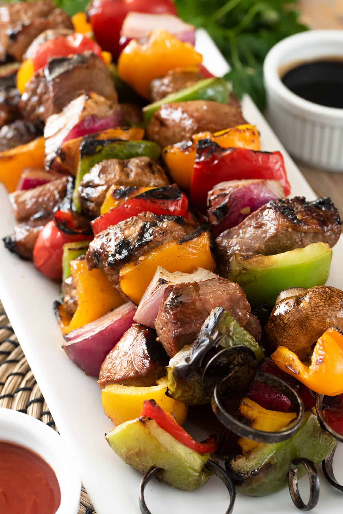 Side view of steak kabobs on metal skewers with peppers, mushrooms, and onions. Sauces on the side.