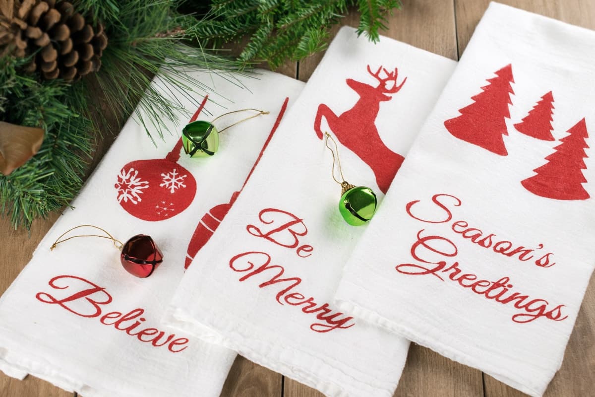 Three white tea towels with red Christmas themed stencils spread on wooden surface with garland, pine cones, and ornaments.