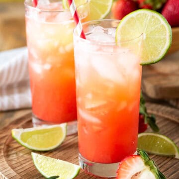 Two glasses of strawberry limeade with lime garnish on a wood background.