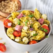 A bowl of pesto tortellini pasta served cold with mozzarella, salami, and tomatoes on top.