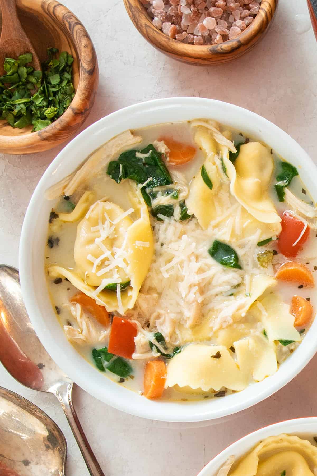 Overhead of a bowl of Chicken tortellini soup recipe in a bowl with herbs in small bowls around it.