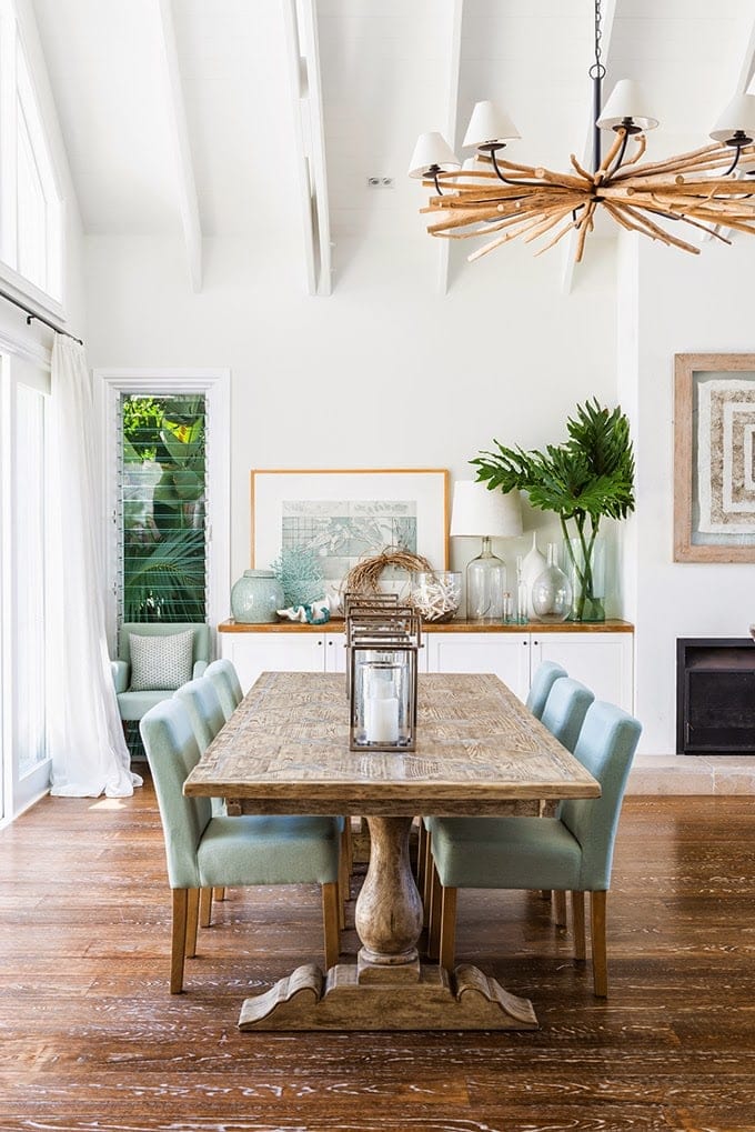 Dining room with white walls and white vaulted ceiling with wood dining table and teal upholstered chairs, topped with lanterns. Cabinets topped with framed picture, plant, and glass vases.