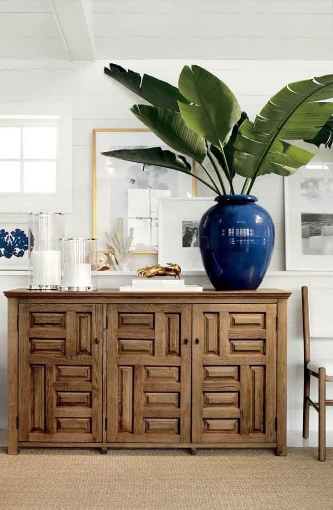 Geometric wooden credenza with large plant in blue ceramic pot, white candles in glass vessels in front of white paneled wall with framed pictures.