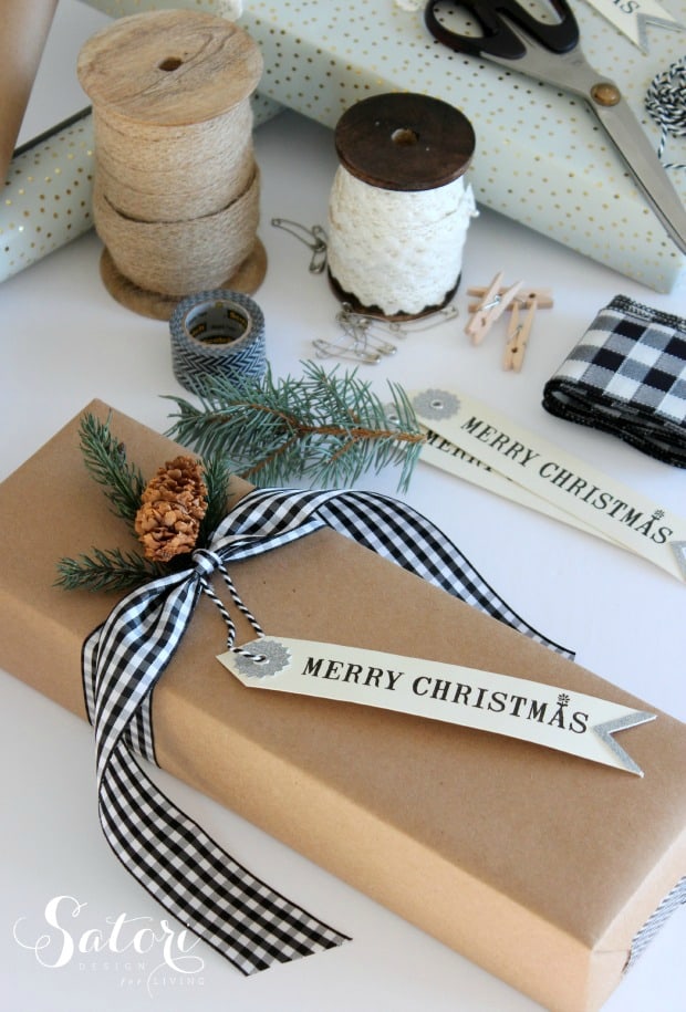 Rectangular package wrapped in craft paper tied with buffalo plaid bow with canvas gift tag and a pine cone.