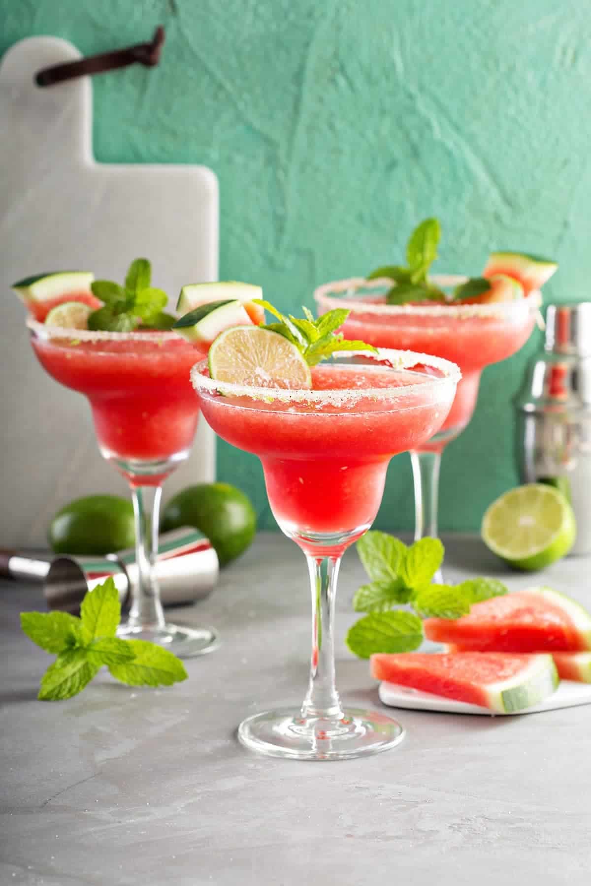 Watermelon margaritas with sugar rim and lime & mint garnish. Cocktail shaker and additional garnishes laid around.