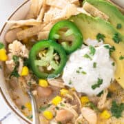 Top down view of a bowl of white chicken chili garnished with avocado, sour cream, jalapeno, and tortilla strips.