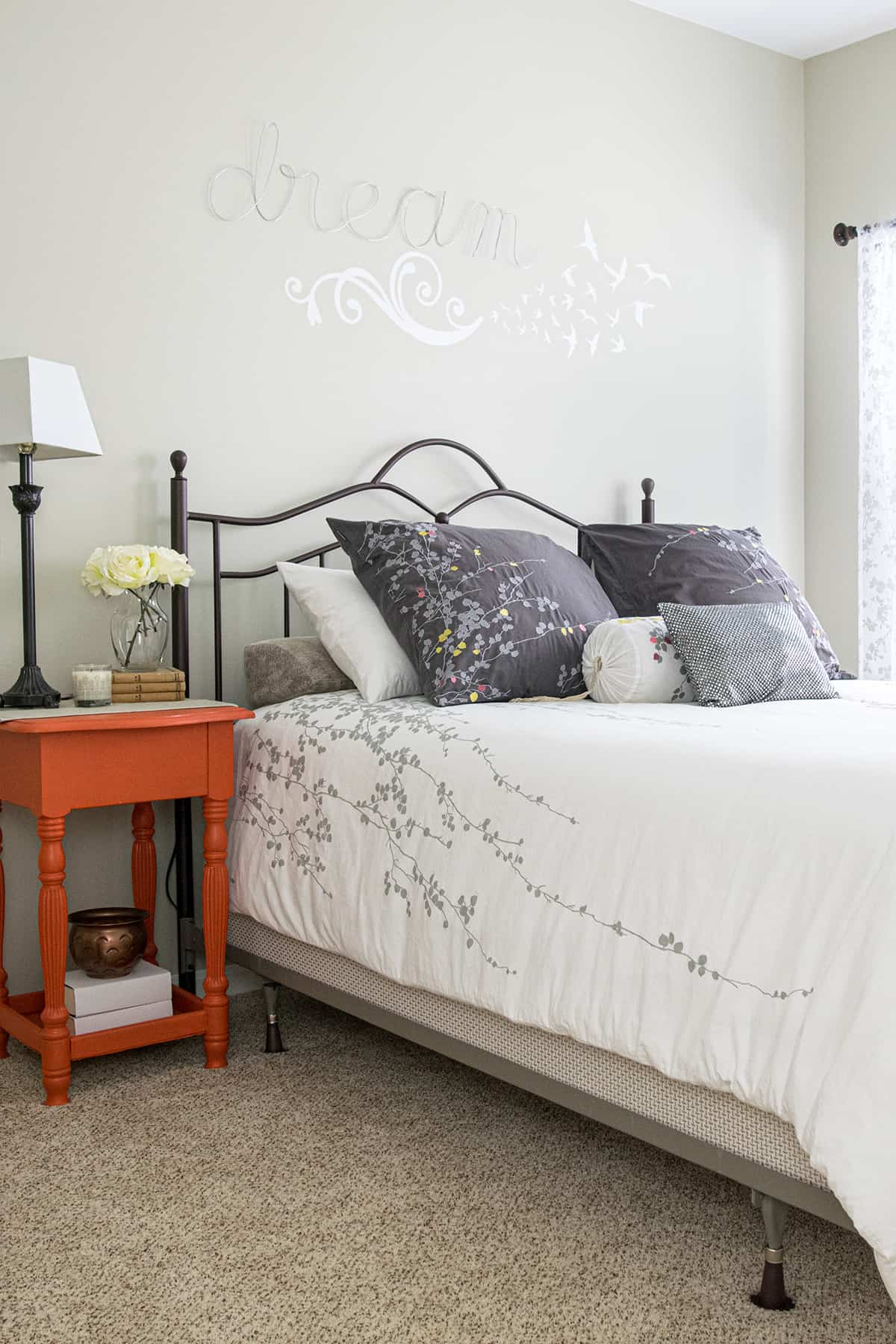 Neutral bedroom with white floral comforter, orange side table, and art over the bed.