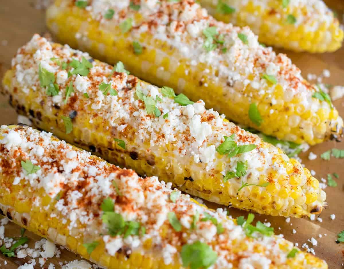 Elotes with cotija cheese, cilantro, garlic-lime sauce, cilantro, and chili powder presented on corn cobs.